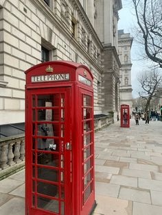 two red telephone booths sitting on the side of a street next to a building with people walking by