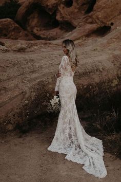 a woman standing in the desert wearing a long sleeved wedding dress and holding a bouquet