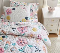 a white bed topped with lots of pillows and quilted bedspread next to a night stand