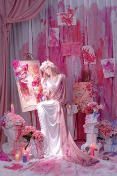 a woman dressed in white and pink sitting next to a painting on a easel