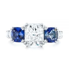 102972 Platinum Ring   2 Blue Sapphires - .91 ctw   10 Diamonds - .98 ctw   Clarity: VS2 - Color: F-G    Joseph Jewelry. This dazzling engagement ring features a radiant cut diamond in the center, with a cushion shaped blue sapphire on either side, and shared prong set diamonds down the top of the platinum Euroshank. It was created for a couple from Issaquah, WA, who already had all three stones they wanted to use for the main setting, as well as a clear vision for what they wanted the overall p Blue Diamond Ring With Round Cut, Blue Round Cut Diamond Ring, Blue Radiant Cut Three-stone Ring, Three Stone Sapphire Ring With Radiant Cut Diamond, Blue Three Stone Round Cut Diamond Ring, Sapphire Cushion Cut Diamond Ring With Center Stone, Sapphire Cushion Cut Ring With Prong Setting, Blue Sapphire Ring In Radiant Cut Platinum, Blue Diamond Ring With Brilliant Cut Lab-created Sapphire