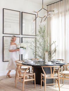 a woman walking past a dining room table with chairs and pictures on the wall behind it