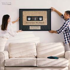 a man and woman holding up a framed audio tape recorder on a wall above a white couch