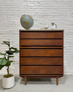 a wooden dresser sitting next to a potted plant on top of a white brick wall