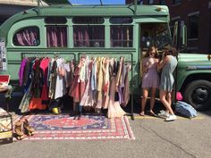 two women standing in front of a green bus with clothes hanging on it's rack