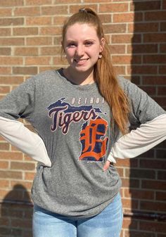 a girl with her hands on her hips standing in front of a brick wall wearing a detroit tigers t - shirt