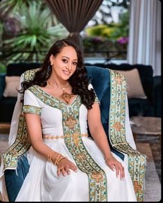 a woman sitting in a chair wearing a white dress and gold jewelry on her neck