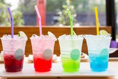 three different colored drinks with straws on a white tray in front of a window