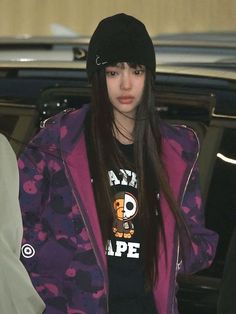 a girl with long hair wearing a purple jacket and black beanie is standing in front of a car