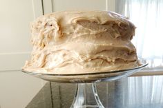 a frosted cake sitting on top of a glass platter next to a window