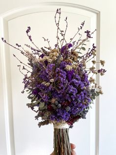 a hand holding a vase with purple flowers in it and greenery on the inside