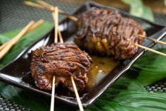 two pieces of meat on skewers sitting on a black plate with green leaves