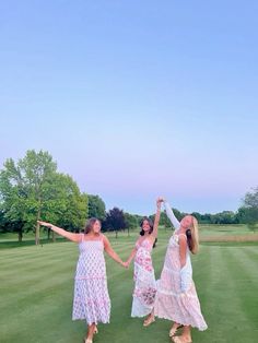 three women in dresses holding hands and dancing on a green golf course with trees in the background