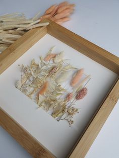 an arrangement of dried flowers in a wooden frame on a white surface with two stalks of grass behind it