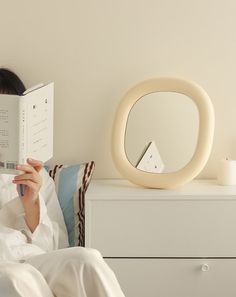 a woman reading a book while sitting on a bed in front of a round mirror