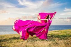 a woman in a pink dress is standing on the grass by the ocean with her arms outstretched