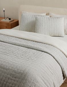 a white bed with two pillows on top of it and a night stand in the background