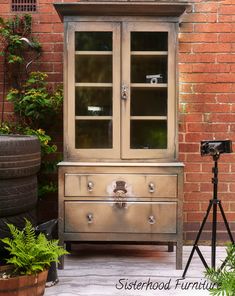 an old china cabinet with glass doors in front of a brick wall and potted plants