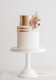 a three tiered white and gold wedding cake with flowers on the top, sitting on a pedestal