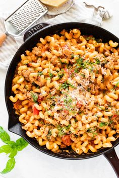 a skillet filled with pasta and meat on top of a white table next to some parmesan cheese