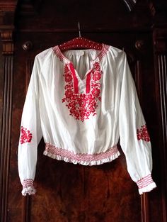 Beautiful 1980s Vintage Hungarian embroidered blouse. White airy fabric with embroidered flowers and vines in a vibrant shade of red on the front and the sleeves. Red zic-zac stitching around the neckline and cuffs. Split neckline. Wide raglan sleeves. Smocked elasticated waist with red zic-zac stitching. Press button closure on the waist. Traditional Hungarian blouses will always be a staple for Bohemians. Looks great with a a pair of highwaist flared jeans and brown clogs! BRAND: No brand labe Folk Style Embroidered Tunic Top For Spring, Long Sleeve Embroidered Top For Spring Festival, Folk Blouse With Embroidered Sleeves For Spring, Peasant Style Embroidered Top With Geometric Embroidery For Spring, Traditional White Peasant Top For Fall, Peasant Style Long Sleeve Embroidered Top For Festivals, Fitted Traditional Embroidered Top For Spring, Folk Style Peasant Top With Embroidered Sleeves For Festival, Long Sleeve Peasant Top With Multicolor Embroidery For Festival