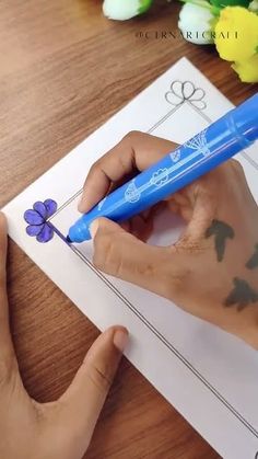 a person is drawing on paper with a blue pen and some flowers in the background