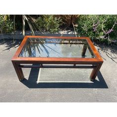 a wooden table with glass top sitting in the middle of a parking lot next to plants