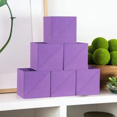 purple cubes are stacked on top of each other in front of green balls and a potted plant