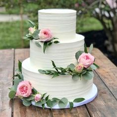 a three tiered white cake with pink flowers on it sitting on a wooden table