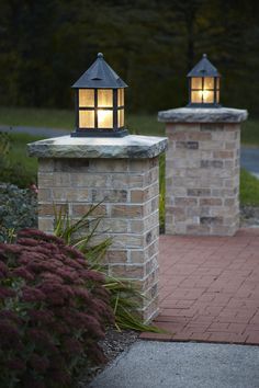 two lights on top of brick pillars in the middle of a walkway with flowers and bushes