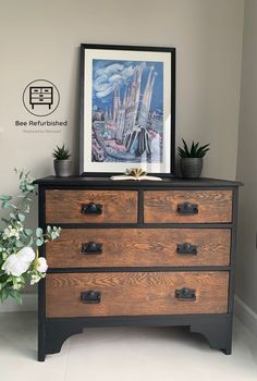 a wooden dresser with two drawers and a painting on the wall above it next to a potted plant