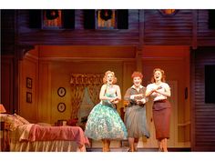 three women in dresses stand on stage with their arms around each other as they laugh