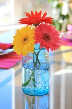 three colorful flowers in a blue mason jar on a glass table with pink and yellow daisies