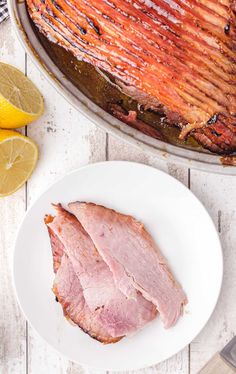 ham on a white plate next to sliced lemons and a large roasting pan