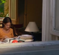 a woman sitting at a table with books and a lamp in front of her on the window sill