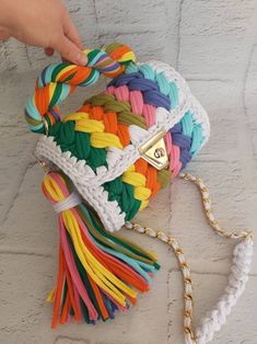 a hand holding a multicolored purse on top of a white brick wall next to a chain