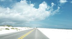 an empty road on the beach with white sand and blue skies in the background, under a partly cloudy sky