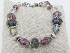 "This ARTISAN GLASS (Lampwork) BRACELET features 3 BOROSILICATE GLASS BEADS in shades of purple and green. The Boro glass beads are roughly 8x15mm and are spaced throughout the bracelet. There are also 10x14mm crystal tapered faceted ovals in purple and 8x12mm faceted crystal rondelles in the same purple; 5x8mm silver spacer beads, along with assorted clear and lavender faceted crystals. There are 7 assorted drops on the chain, inclding lampwork rondelles, Swarovski crystals, other glass, crysta Useable Gifts, Lampwork Bracelets, Beautiful Beaded Necklaces, Green Bracelet, Farmers Market Bag, Glass Lampwork, Fitness Bracelet, Glass Bracelet, Purple And Green