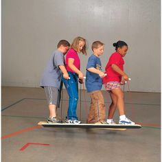 four children are standing on a skateboard in an indoor gym with ropes attached to the board