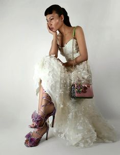 a woman in a white dress is holding a purse and posing for the camera with her hand on her face