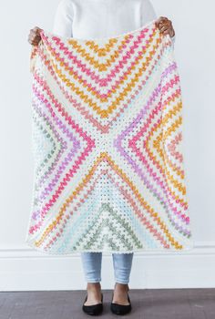 a woman holding up a multicolored crocheted blanket in front of a white wall