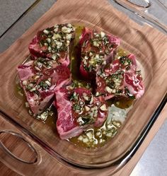 some meat is sitting in a glass dish on a wooden tray with olives and seasoning