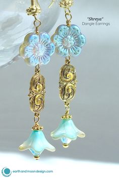 These striking dangle earrings are full of alluring color and shimmer. Lovely bell flowers in dreamy aqua-blue hue with a delicate gold finish and are made of Czech glass and donned with gold-toned pewter embellishments and finished with an aqua-blue Swarovski crystal. They are suspended from decorative gold-toned pewter accents and Czech glass daisies that are washed with an iridescent purple-blue-gold finish. Earrings fall 2" from ornate gold-plated brass ear wires. $36 Feminine Drop Earrings For Spring, Nickel-free Czech Glass Flower Dangle Earrings, Nickel-free Czech Glass Dangle Earrings, Nickel-free Czech Glass Flower Jewelry, Delicate Drop Earrings For Spring, Elegant Teardrop Earrings For Spring, Feminine Jewelry With Matching Earrings For Spring, Feminine Spring Dangle Earrings, Spring Feminine Dangle Jewelry