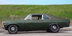 an old green car is parked in front of a fence and grassy hill behind it