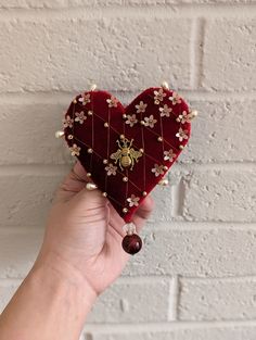 a hand holding a red heart shaped box with pearls and beads on the sides, against a white brick wall