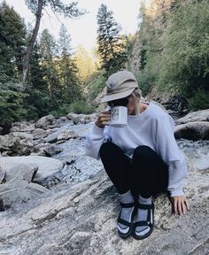 a woman sitting on top of a rock next to a river holding a coffee cup