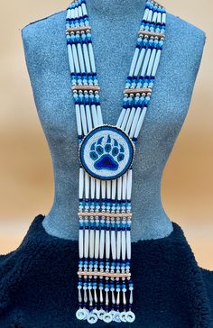 Bone hairpipes and crystal beads are often seen as part of traditional Native American dancers.  This modified breastplate  has been created to proudly proclaim that this is a good day to be Indigenous . It is designed to be worn not only in the dance circle, but in mainstream life, showing your Native pride and fashion sense.   The necklace is 10 vertical strands of bone hairpipes, nickel beads and 8mm sparking, faceted, fire-polished glass crystal beads in the colors of navy blue, turquoise blue and white frost The centerpiece is a beaded rosette of a Bear paw in the same blue colors on a white background. At the bottom of breastplate, pewter and rhinestone pendants  add their music to your movements. The breastplate comes with matching earrings on hypoallergenic lever back wires featuri Medallion Necklace Beaded, Native American Medallions, Powwow Regalia Jingle Dress, Beaded Medallion Native American, Native Medallion, Indigenous Jewelry, Indigenous Fashion, Native American Dance, American Dance