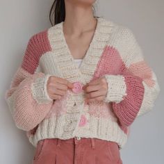 a woman wearing a pink and white cardigan with buttons on the front, standing in front of a wall