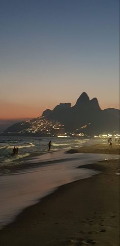 people are walking on the beach at night with mountains in the background and lights reflecting off the water