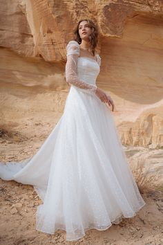 a woman in a white wedding dress standing on the sand with her arms behind her back